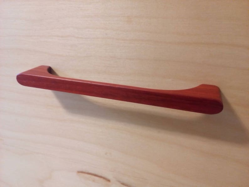 Pode incluir: Um puxador de arm&aacute;rio de madeira castanho-avermelhado com uma pega curva. O puxador &eacute; montado numa superf&iacute;cie de madeira clara. A pega tem aproximadamente 25 cm de comprimento.