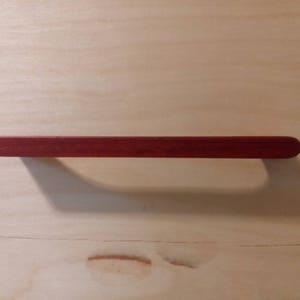 Pode incluir: Puxador de arm&aacute;rio de madeira maci&ccedil;a, vermelho escuro, com formato retangular e extremidades arredondadas. O puxador est&aacute; sobre uma superf&iacute;cie de madeira clara, projetando uma sombra.