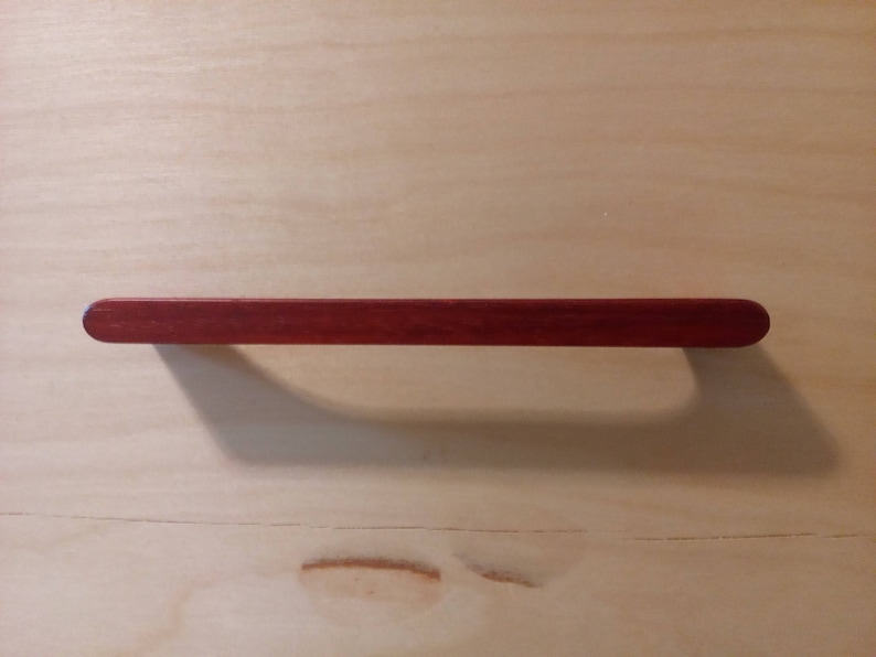 Pode incluir: Puxador de arm&aacute;rio de madeira maci&ccedil;a, vermelho escuro, com formato retangular e extremidades arredondadas. O puxador est&aacute; sobre uma superf&iacute;cie de madeira clara, projetando uma sombra.