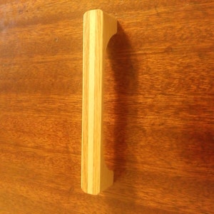 May include: A light-colored wooden cabinet pull with a rectangular shape and a central light-colored stripe. The handle is made of multiple pieces of wood and is designed for easy gripping. The pull is set against a dark wood surface.