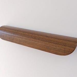 May include: A wooden cabinet pull with a curved, ergonomic design. The pull is made of a rich, dark brown wood, showcasing the natural grain. The handle is approximately 8 inches long.