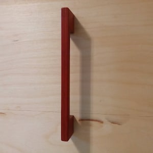 May include: A red wooden cabinet pull handle. The handle is rectangular with a flat, vertical design. The handle is mounted on a light-colored wooden surface.