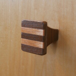May include: A rectangular wooden cabinet knob with a dark brown and light brown striped design. The knob is attached to a light-colored wooden cabinet door. The knob's design is a mix of dark and light wood tones.