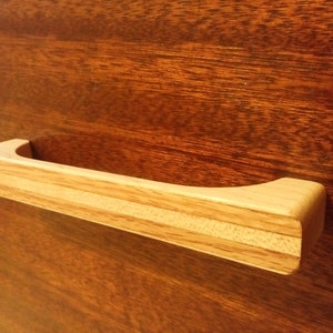 Peut inclure: Une poignée de tiroir en bois clair de forme rectangulaire avec une poignée incurvée. La poignée est faite de bois en couches, avec un bois plus clair au centre et un bois plus foncé sur les côtés. Elle est montée sur une surface en bois brun foncé.