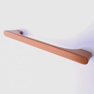 May include: A wooden cabinet pull with a light brown finish. The handle is rectangular with rounded edges and a curved design on each end. The pull is approximately 12 inches long.