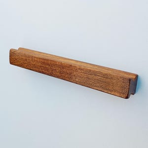 May include: A wooden drawer pull with a rectangular shape and a slightly raised center section. The pull has a natural wood finish and a slightly rough texture.