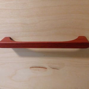 Pode incluir: Um puxador de arm&aacute;rio de madeira castanho-avermelhado com um design curvo. A pega &eacute; retangular com uma forma ligeiramente curva e &eacute; montada numa superf&iacute;cie de madeira de cor clara. O puxador tem cerca de 25 cm de comprimento.