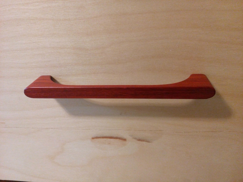 Pode incluir: Um puxador de arm&aacute;rio de madeira castanho-avermelhado com um design curvo. A pega &eacute; retangular com uma forma ligeiramente curva e &eacute; montada numa superf&iacute;cie de madeira de cor clara. O puxador tem cerca de 25 cm de comprimento.