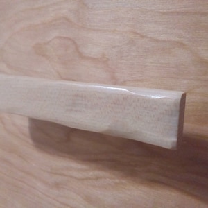 May include: A light-colored, rectangular wooden handle with a rounded end. The handle appears to be smooth and unfinished, with visible wood grain. The handle is set against a light-colored wooden surface.