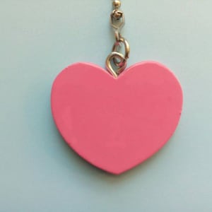 Puede incluir: Un colgante rosa en forma de corazón suspendido de una cadena plateada. El colgante es de un color rosa sólido y vibrante, sobre un fondo azul claro. La cadena está unida a un pequeño elemento metálico decorativo.