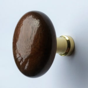 Puede incluir: Un pomo de armario ovalado de madera con un acabado marrón oscuro brillante. El pomo está unido a una base color crema con un anillo de metal dorado. El herraje está diseñado para muebles y armarios.
