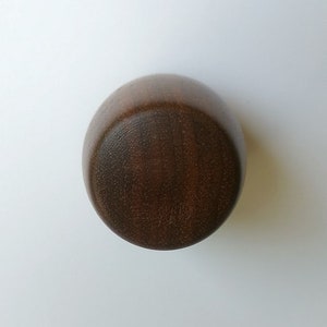 Puede incluir: Un objeto redondo de madera marrón oscuro con una superficie lisa y curvada. Parece un cuenco o recipiente pequeño, posiblemente para uso decorativo o funcional. La veta de la madera es visible, añadiendo textura al diseño.