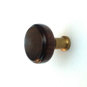 Puede incluir: Perilla de armario redonda de madera marrón oscuro con base de latón. La perilla tiene un acabado liso y pulido y está sobre un fondo blanco. Adecuado para cajones, armarios y otros muebles.