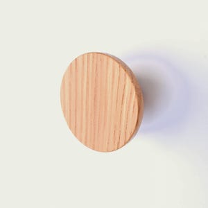 May include: A light brown, wooden, oval-shaped wall hook. The hook is made of wood with visible grain and a natural finish. The hook is mounted on a white surface.