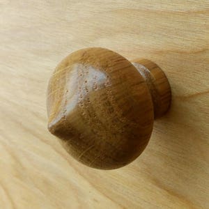 Puede incluir: Primer plano de un tirador de armario de madera con forma redondeada y cónica. El tirador es de madera de color marrón claro y tiene una superficie lisa y pulida. El tirador está fijado a una superficie de madera clara.