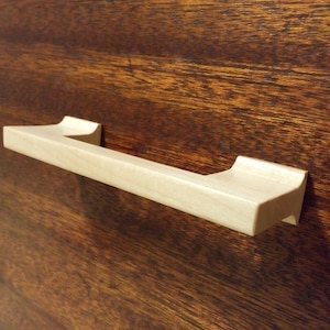 Könnte beinhalten: Ein hellfarbener Holzschrankgriff mit rechteckigem Griff und abgerundeten Kanten. Der Griff ist auf einer dunkelbraunen Holzoberfläche montiert, was einen Kontrast in Farbe und Textur erzeugt. Die Hardware ist für Möbel konzipiert.