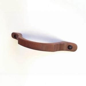 May include: A curved wooden drawer pull with a smooth, medium-brown finish. The handle features a rounded, ergonomic shape and is secured at both ends by dark, flat-head metal screws that are visible on the mounting plates of the hardware.