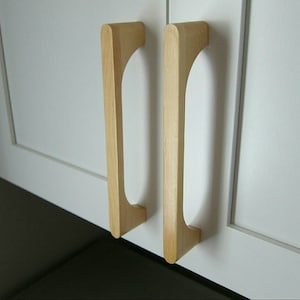 Puede incluir: Dos tiradores de armario de madera con forma rectangular redondeada y una ligera curva en la parte superior. Los tiradores son de color madera claro y están montados en puertas de armario blancas.