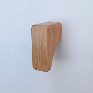 May include: A wooden cabinet knob with a light brown finish. The knob is rectangular with rounded edges and a slight curve on one side.
