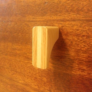 Könnte beinhalten: Ein Holzschrankgriff mit geschwungenem Design. Der Griff besteht aus hellem Holz mit vertikalen Maserungsmustern, befestigt an einer dunkelbraunen Holzoberfläche. Der Griff ist etwa 7,6 cm lang.