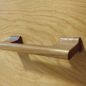 May include: A light brown wooden cabinet pull with a rectangular handle and curved ends. The pull is mounted on a light brown wooden surface, showcasing the natural wood grain.