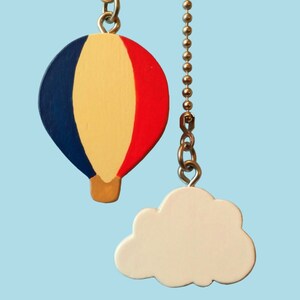 Pode incluir: Um puxador de ventilador de teto lúdico com um balão de ar quente pintado à mão em vermelho, amarelo e azul, com um puxador em forma de nuvem branca. O balão é suspenso por uma corrente de metal sobre um fundo azul claro.