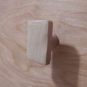Puede incluir: Un pomo de armario rectangular de madera clara con una base redondeada. El pomo está fijado a una superficie de madera clara, con la veta de la madera visible.