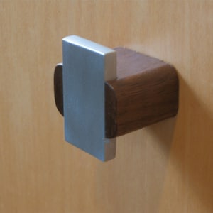 May include: A wooden cabinet pull with a rectangular, brushed metal handle. The pull is mounted on a light-colored wooden surface. The wooden portion is a dark brown color.