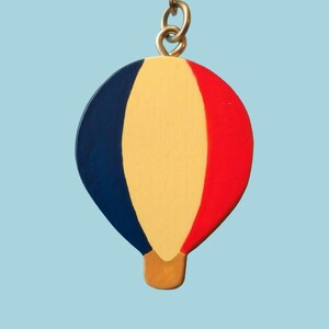 Puede incluir: Un llavero de globo aerostático azul, amarillo y rojo. El globo está hecho de madera y tiene una base dorada.