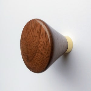 May include: A wooden cabinet knob with a conical shape. The knob is made of dark brown wood with a visible grain pattern. The base of the knob is a light cream color, and it is mounted on a white surface.