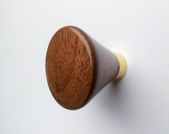 May include: A wooden cabinet knob with a conical shape. The knob is made of dark brown wood with a visible grain pattern. The base of the knob is a light cream color, and it is mounted on a white surface.