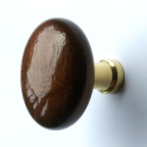 May include: A round cabinet knob featuring a polished dark brown wooden head with a visible grain pattern. The knob is attached to a cylindrical base made of brushed gold-toned metal, which connects to a circular mounting plate at the rear.