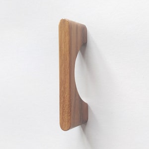 May include: A wooden cabinet door pull with a rounded rectangular shape and a semi-circular cutout. The pull is made of a light brown wood with a natural grain pattern.