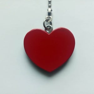 May include: A bright red heart-shaped charm, suspended from a silver chain. The heart is smooth and glossy, with a solid color. The background is a plain white surface. This charm could be used as a pendant or a decorative pull.