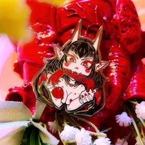 May include: A detailed enamel pin featuring a stylized anime-inspired character with red eyes, black hair, and pointed ears. The pin includes the text "Bite the hand that feeds" on a red banner. The character is holding a red apple.