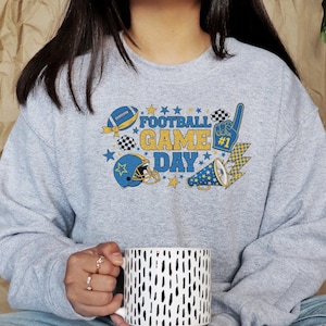 May include: A light grey sweatshirt featuring a football-themed design. The graphic includes a football, helmet, megaphone, and the words "FOOTBALL GAME DAY" in blue and yellow. A white and black patterned mug is held in the hands.