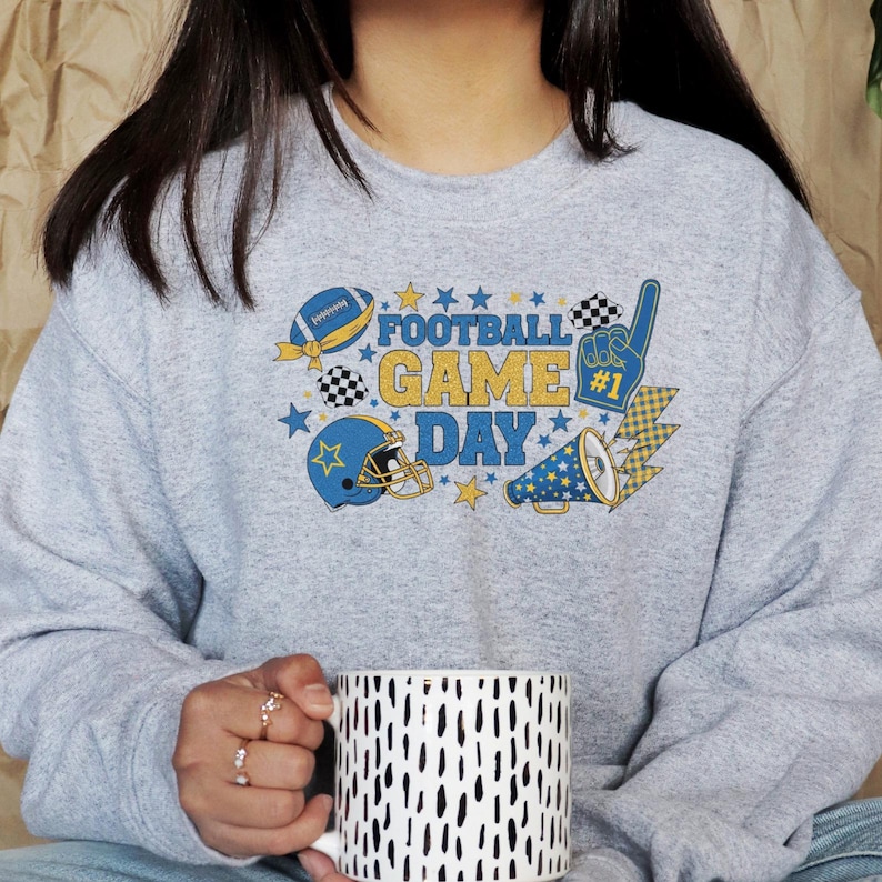 May include: A light grey sweatshirt featuring a football-themed design. The graphic includes a football, helmet, megaphone, and the words "FOOTBALL GAME DAY" in blue and yellow. A white and black patterned mug is held in the hands.