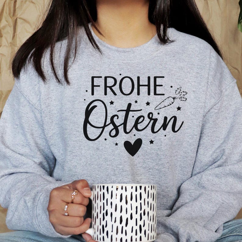 K&ouml;nnte beinhalten: Ein hellgraues Sweatshirt mit dem schwarzen Schriftzug "FROHE OSTERN", einer Karottenillustration und einem Herz. Eine wei&szlig;e Tasse mit schwarzen vertikalen Linien wird in den H&auml;nden gehalten.