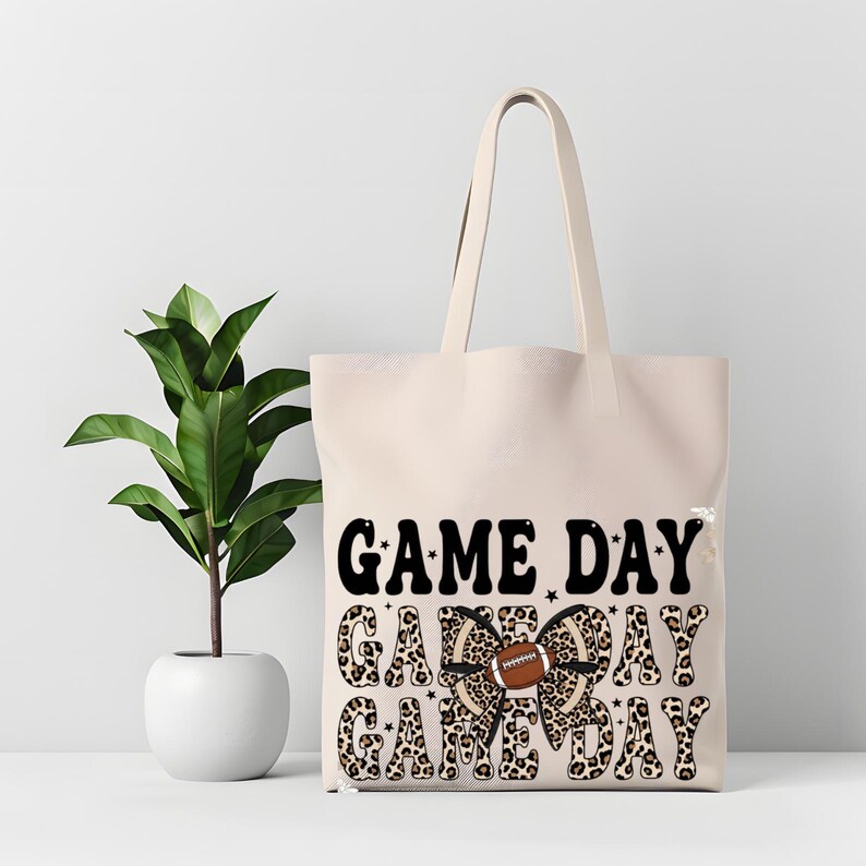 May include: A cream-coloured tote bag featuring the text "GAME DAY" in black, with a leopard print bow and a football graphic. The bag has long handles and is made of a durable material.
