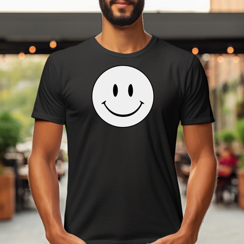 May include: Black t-shirt featuring a large white smiley face graphic. The smiley face has two black dots for eyes and a curved black line for a mouth. The shirt is a classic crew neck style.