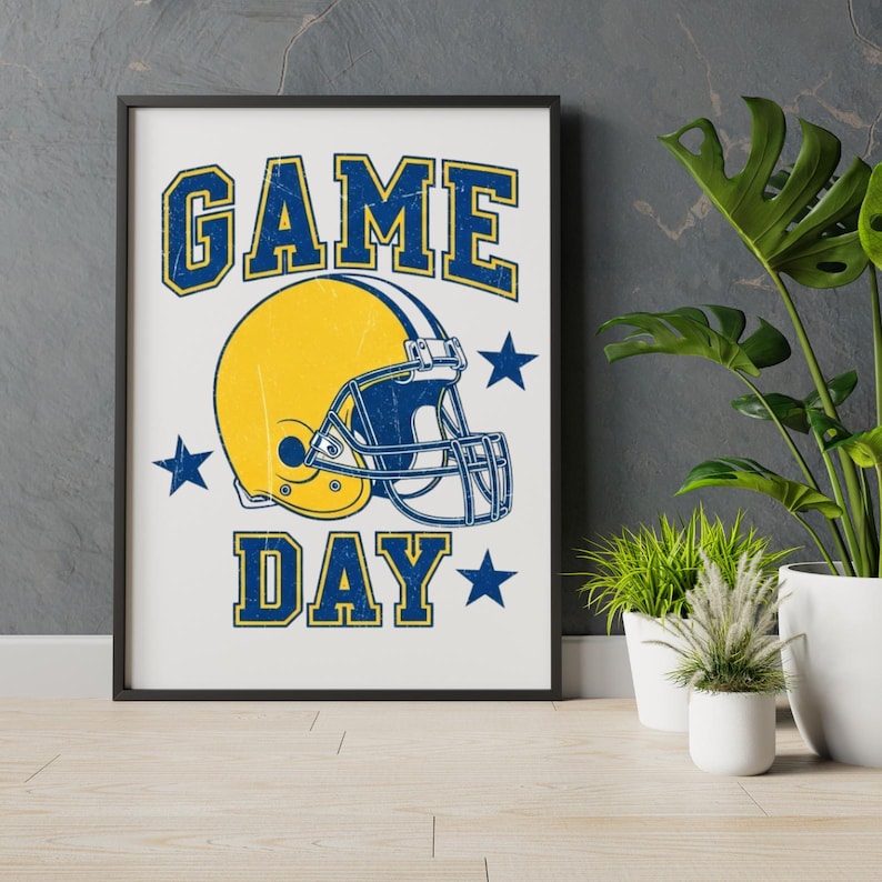May include: Framed print with a yellow and blue football helmet and the words "GAME DAY". The text and helmet have a distressed effect. Blue stars surround the helmet. The print is in a black frame, against a grey wall.