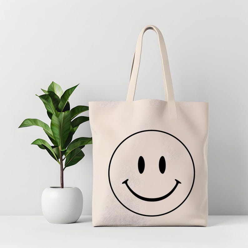 May include: A beige tote bag featuring a large black smiley face. The bag has a simple design with a rectangular shape and two handles. A small potted plant with green leaves is to the left of the bag.