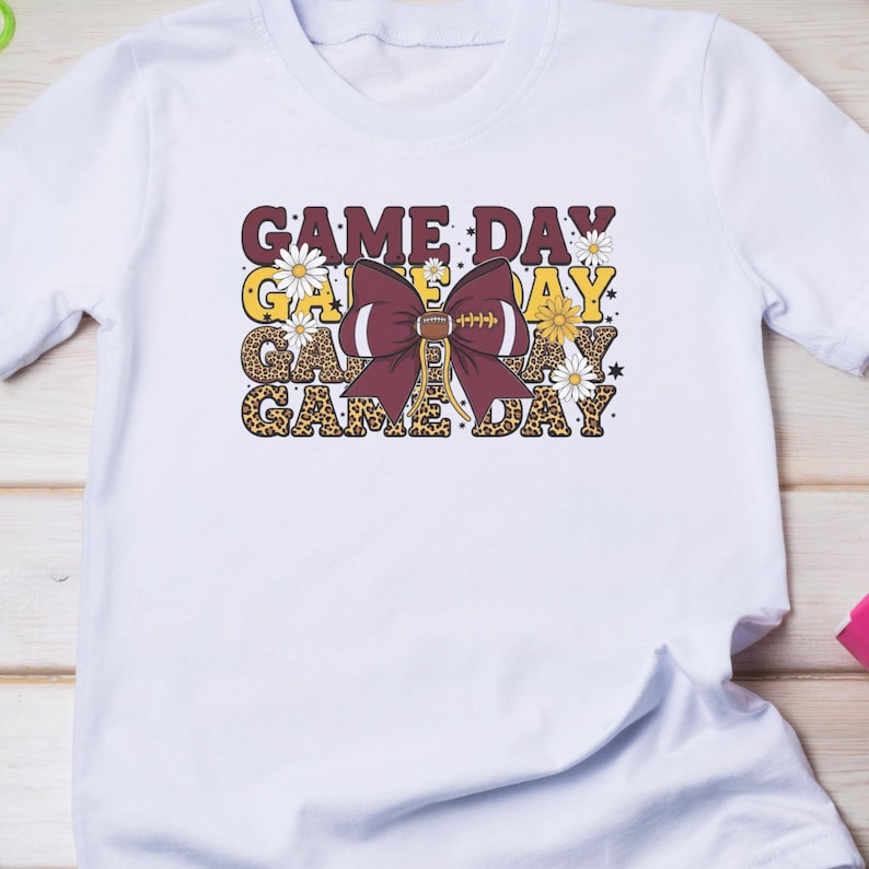 May include: White t-shirt with the words "GAME DAY" in a retro font. The design includes a football-shaped bow with football lace detail, daisies, and a leopard print pattern. A fun and stylish tee for game day.
