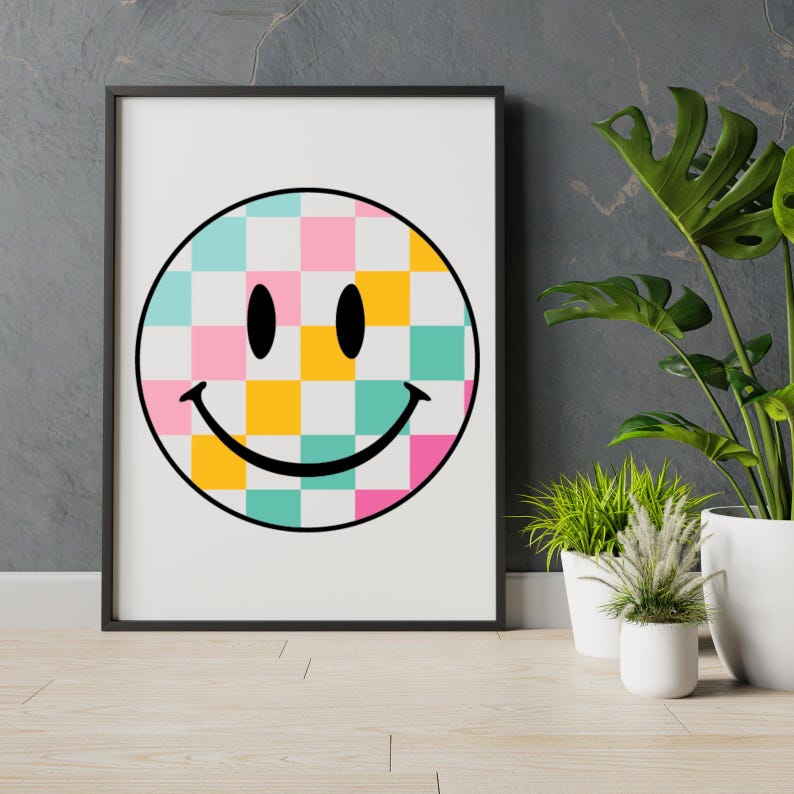 May include: A framed print featuring a smiley face design. The smiley face has a black outline and a checkered pattern in shades of pink, yellow, and teal. The print is in a black frame and is displayed against a gray wall.