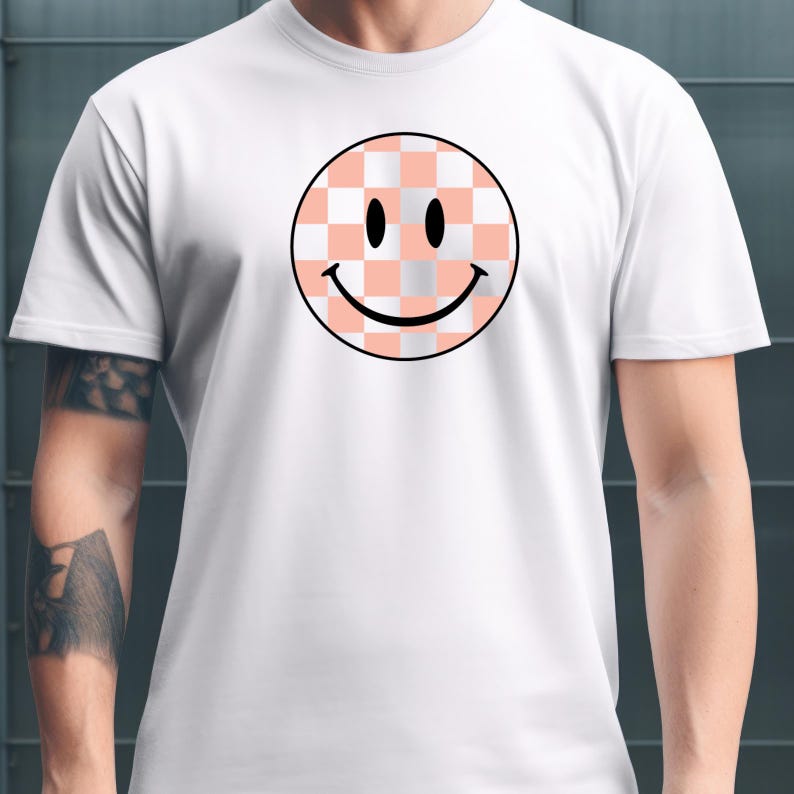 May include: White t-shirt featuring a large, circular smiley face design. The smiley face has a black outline and a checkered pattern in shades of peach and white. The eyes are black dots, and the mouth is a curved black line.