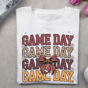 May include: White t-shirt featuring the text "GAME DAY" in a layered design. The top and bottom layers are maroon and orange, with a leopard print middle layer. A football with a maroon bow is in the center.