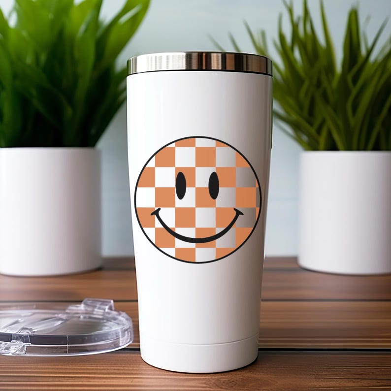 May include: White stainless steel tumbler featuring a smiley face design. The smiley face has a checkered pattern in orange and white. The tumbler has a silver rim and a clear lid. The tumbler is approximately 8 inches tall.