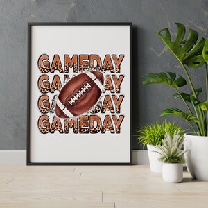 May include: Framed print with the word "GAMEDAY" in a stacked design and a football in the center. The text has a leopard print pattern, and the football is brown with white stripes. The print is in a black frame.