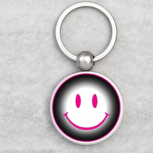 May include: A round keychain with a smiley face design. The face features a white smile and two pink eyes on a black and white gradient background, framed by a pink border. The keychain has a silver metal ring.