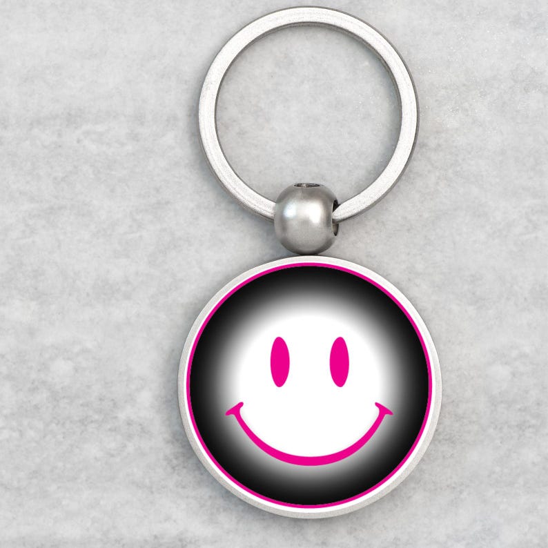 May include: A round keychain with a smiley face design. The face features a white smile and two pink eyes on a black and white gradient background, framed by a pink border. The keychain has a silver metal ring.